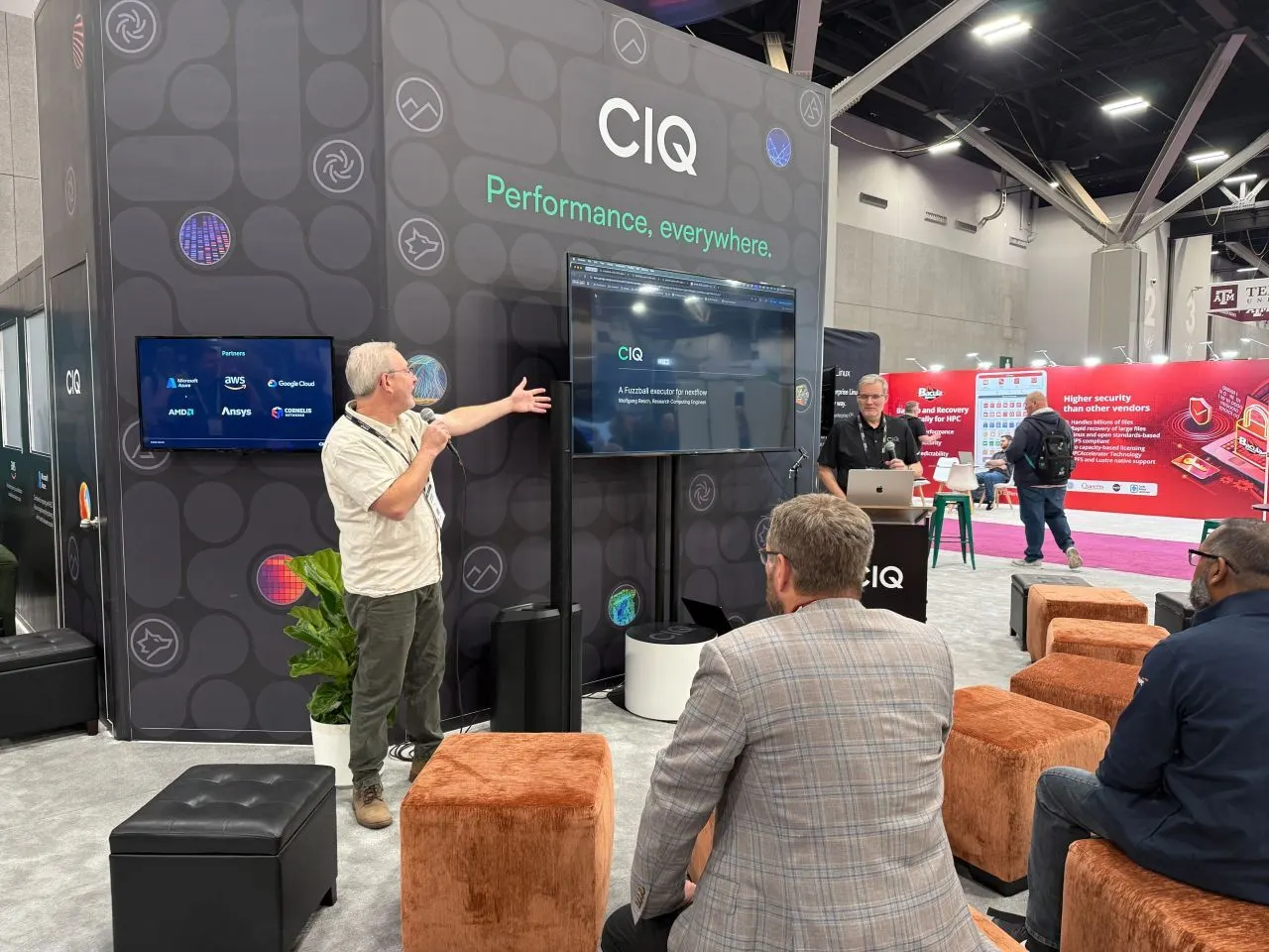 A presenter gesturing toward a screen while visitors sit on ottomans at the CIQ booth